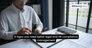 HR compliance management professional reviewing legal and HR compliance documents at desk with Savvy HRMS software visible in background